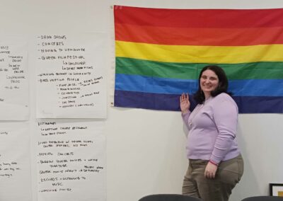 A woman with dark brown hair stands in front of a rainbow pride flag. She is wearing a light purple sweater and brown pants, and is smiling. Next to her are four large sheets of paper, with a long list of suggested ways to connect with queer community.