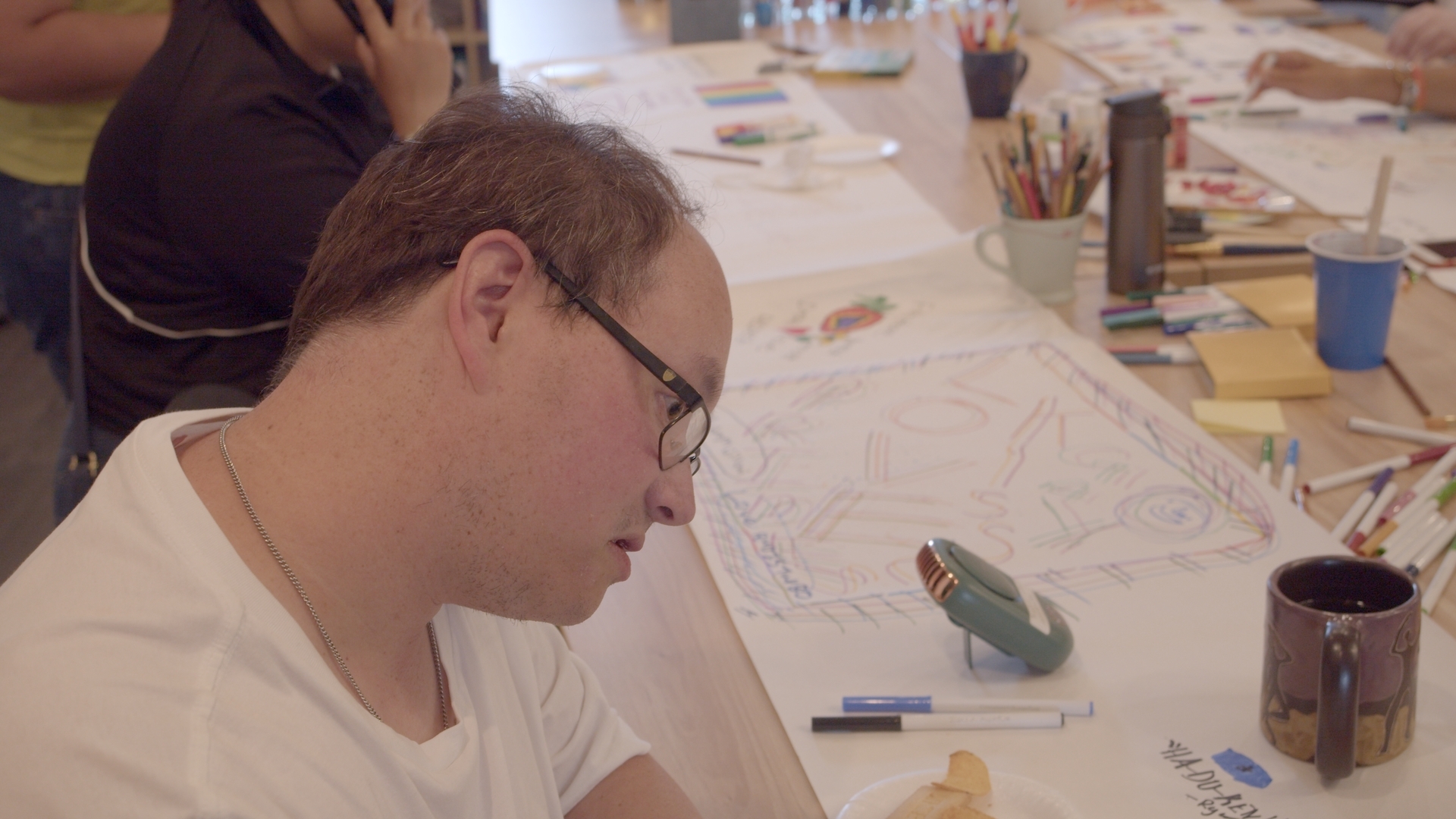 [ID: a person decorating a banner which is on a large table, there is many different colored markers, and other people surrounding the table as well.]